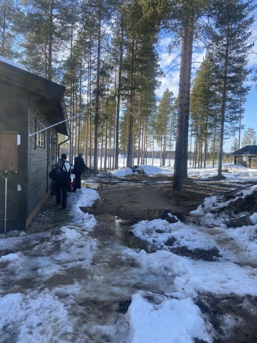 Två personer vid en stuga i snöigt skogslandskap med smältande snö och bar mark.