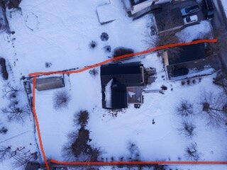 Flygfoto av hus och träd i snö med framträdande orange linje, antagligen illustration av planerad väg eller område.