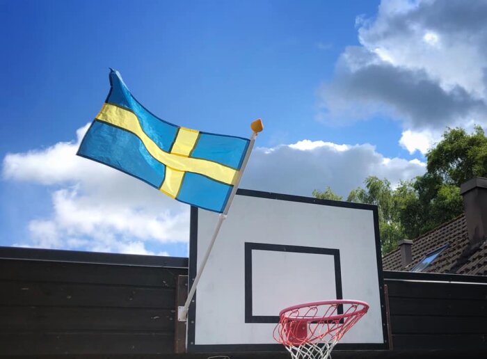 Svensk flagga vajar, bakom basketkorg, blå himmel, vita moln, dagtid.