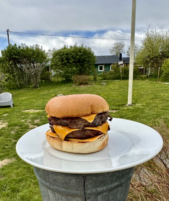 Dubbel cheeseburgare på ett vitt fat utomhus med grönområde och hus i bakgrunden. Cloudy sky.