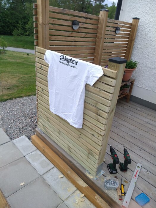 Vit t-shirt hänger på trästaket, verktyg och byggmaterial syns i förgrunden, utomhusmiljö.