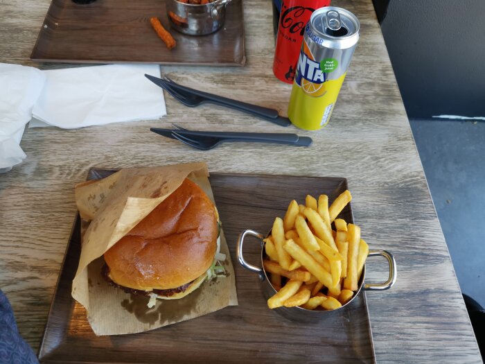 Hamburgare och pommes frites på träbricka, två läskburkar, bestick och servetter på bordet.