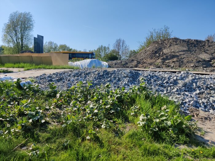 Byggplats med grus, högar av jord, byggnadsmaterial och gröna växter under klarblå himmel.