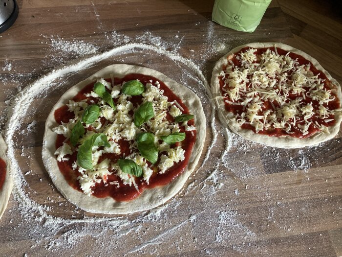 Två otoppade pizzor med tomat, ost, basilika på mjölbestrött träbord. Matlagning hemma.