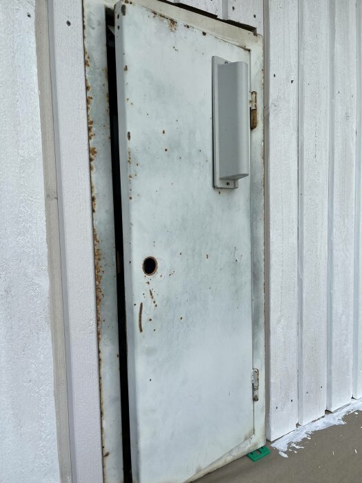 Vit rostig metall dörr med säkerhetsbox, stängd, mot vit trävägg, synlig slitage och korrosion.