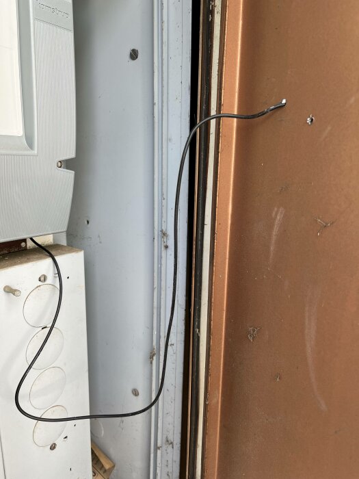 Avskalade svarta kablar hänger löst intill vit apparat och rostig brun dörr.