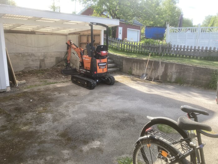 Minigrävare på gårdsplan, cykel i förgrunden, skottkärror och byggmaterial under tak, trästaket, grönska.