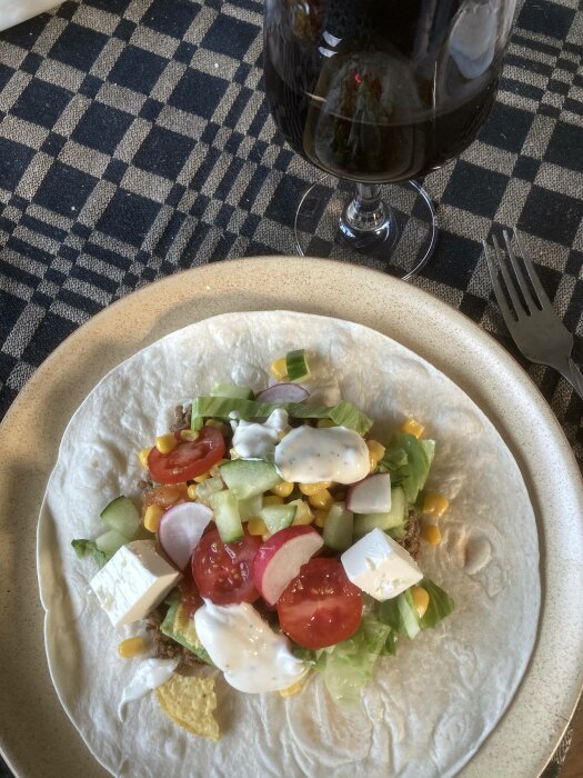 Tortilla med grönsaker, ost, majs, dressing, närbild. Ett glas mörk dryck. Rutig duk. En gaffel. Hemmamiljö.