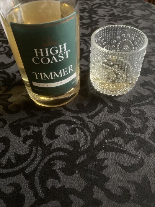 Whiskyflaska märkt 'High Coast TIMMER', tomt glas, mönstrad mörk duk.