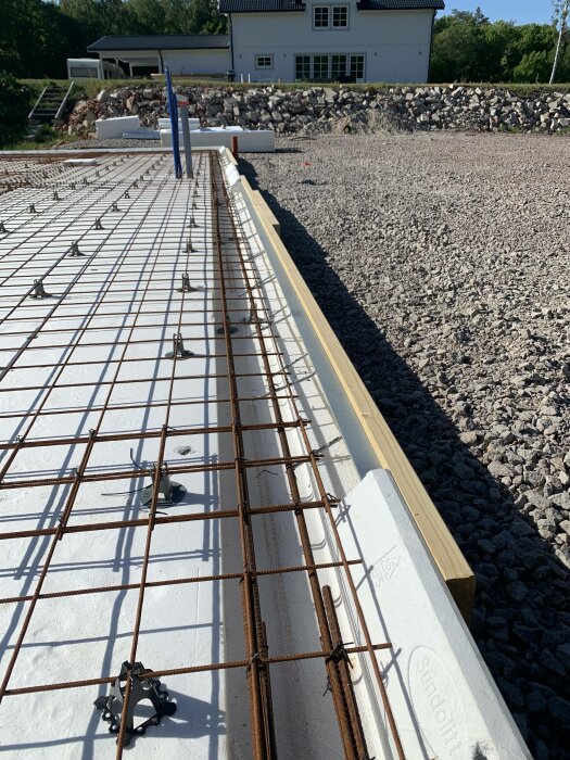 Byggarbetsplats med uppställd armering på isolering inför betonggjutning, klar himmel, hus och grushög i bakgrunden.