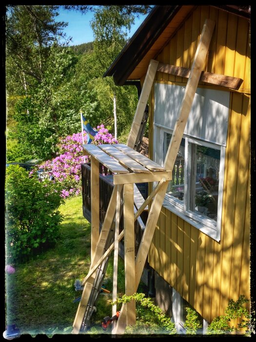 Gulligt gult hus med träställning, blommande buske, svenska flaggan, soligt grönt område, sommarstämning.