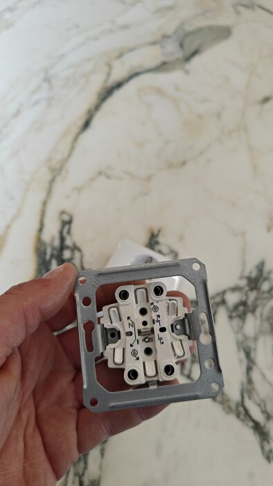 Hand håller elektrisk installationsdosa ovanför marmorbord. Metall, plastdetaljer, skruvanslutningar synliga.