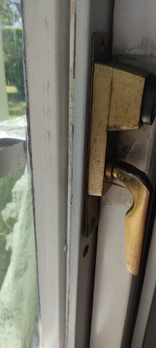 Ett fönsterlås nära karm, slitet, smutsigt, med tydliga tecken på användning, träd syns utanför.