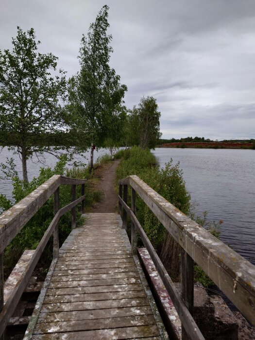 En träbro leder mot en stig längs en sjö, omgiven av gröna träd, under en molnig himmel.