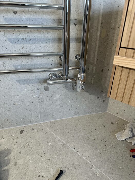 En kromad handdukstork monterad på en betongvägg bredvid ett träpanel, verktyg och trasa på golvet.