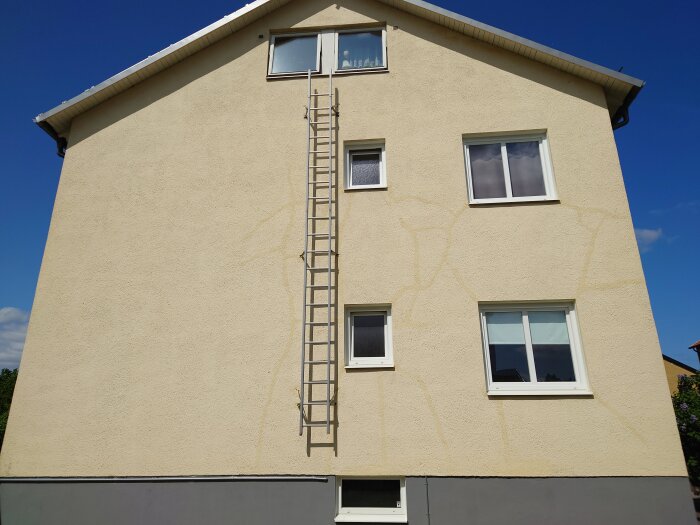 Gult hus, stegar, fönster, blå himmel, synliga reparationsspår på fasaden.