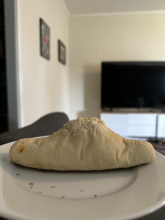 Ett ofärdigt calzone på tallrik i vardagsrum, suddig bakgrund med TV och väggdekorationer.