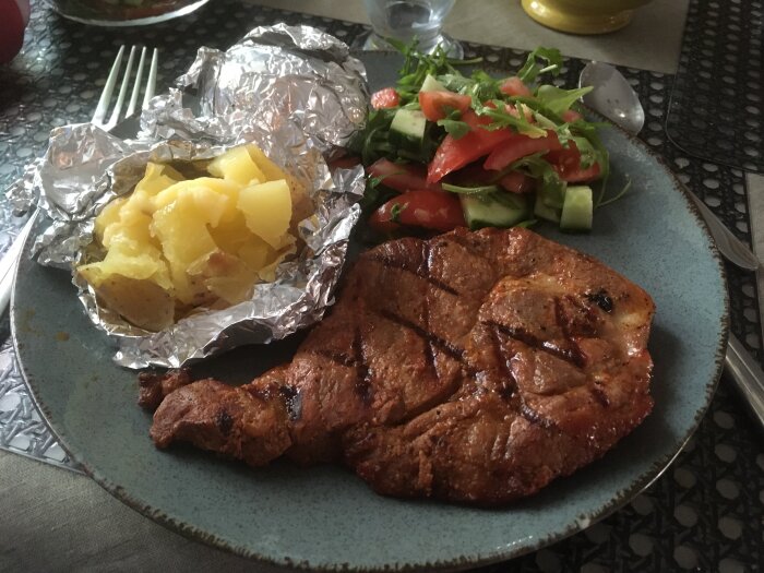 Grillad köttbit, bakade potatisar i folie, färsk sallad, gurka, tomat, bladgrönt på blå tallrik.