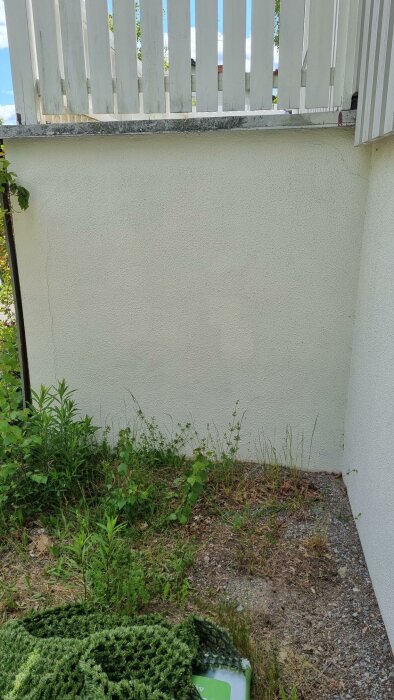 Ett vitmålat staket ovanför en beige vägg, grönska nedanför, gruset och en grön vattenkanna synlig.