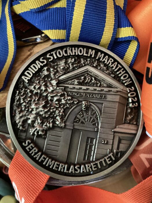 Adidas Stockholm Marathon 2023-medalj med Serafimerlasarettet, blå-gul band, detaljerad prägling.