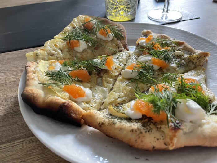 Pizza med löjrom, crème fraiche, rödlök, dill på vit tallrik och glas i bakgrunden.