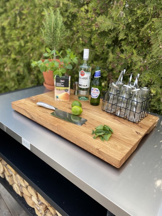 Outdoor bartending setup with alcohol, lime, mint, knife, and glasses on a wooden board.