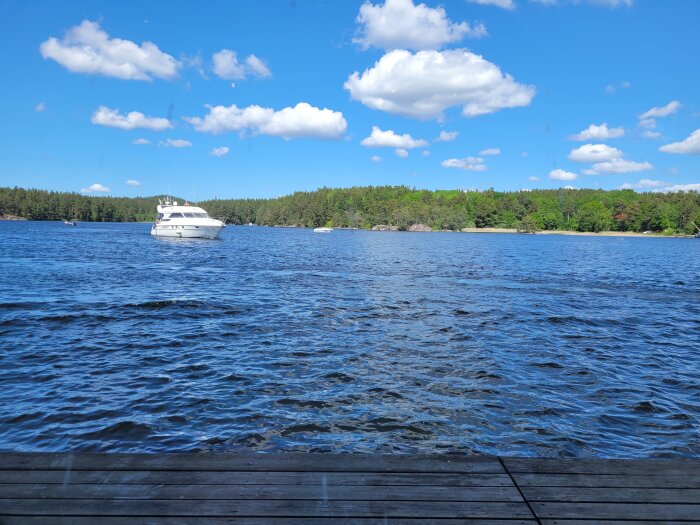 En båt på vattnet, trädäck i förgrunden, skog och blå himmel med moln.