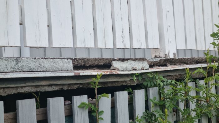 Vitt staket, skadad betonggrund, gröna buskar, tecken på förfall och behov av underhåll.