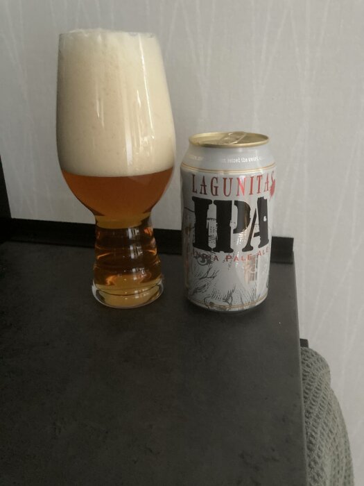 Ölglas med mycket skum bredvid en öppnad Lagunitas IPA-ölburk på en mörk yta.