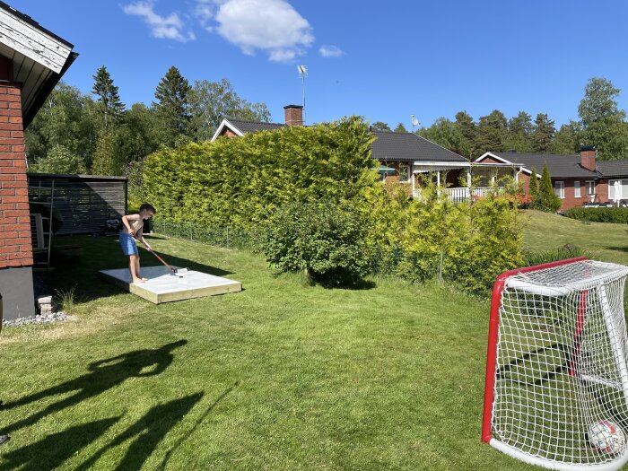 Person målar ett trädeck utomhus, fotbollsmål på gräsmatta, solig dag i villakvarter.