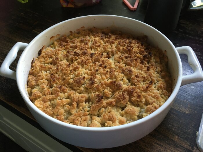 En ugnsform med en gyllenbrun smulpaj, troligen äpple, på ett mörkt bord.