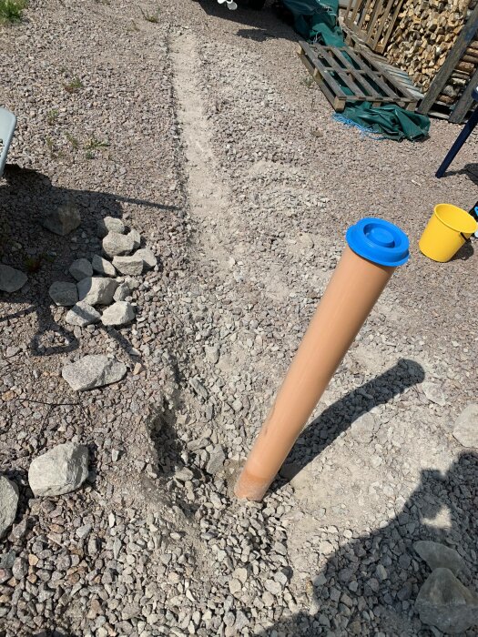 Brun plasttub med blått lock, stenig mark, soligt, skuggor, gelb hink i bakgrunden, utomhus.