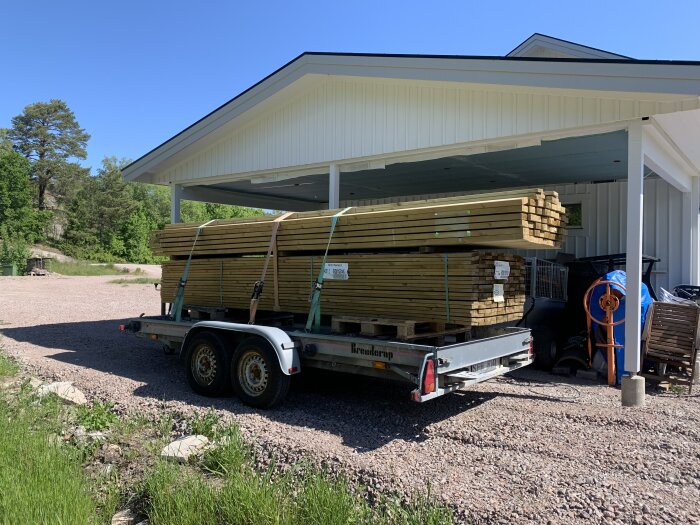 Släpvagn lastad med träbrädor utanför lagerbyggnad, soligt, grusig mark, grönt gräs och träd bakgrund.