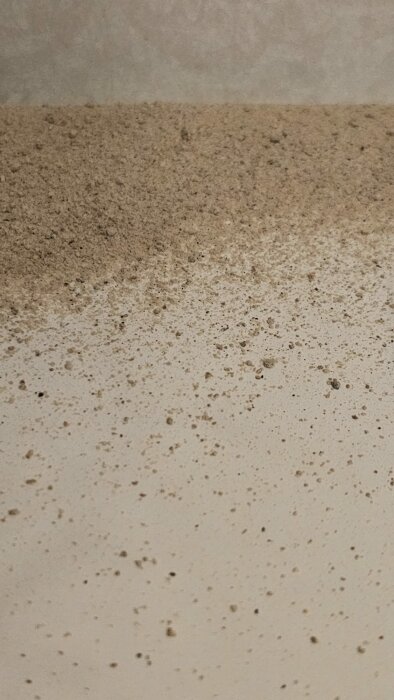 Vägg möter sandfärgat golv täckt av smuts och fläckar. Möjlig fukt- eller mögelskada.
