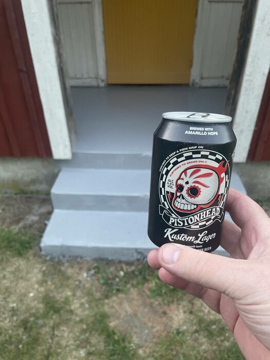 En hand håller en burk öl framför en trappa och en gul dörr.