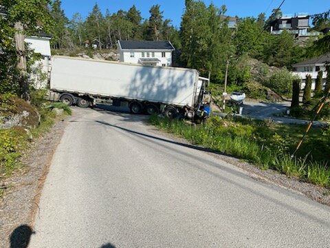 Lastbil blockerar smal landsväg, omgiven av vegetation och hus, solig dag.