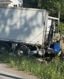 Lastbil med lyftanordning fastnat i diket vid vägkanten, skadat gräs och vegetation under en solig dag.