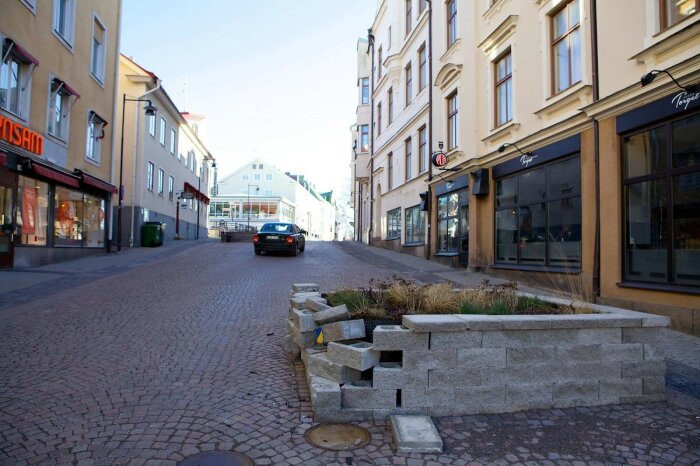 Gata med kullersten, byggnader, plantering, bil, skyltfönster, blå himmel, dagtid, inga människor.