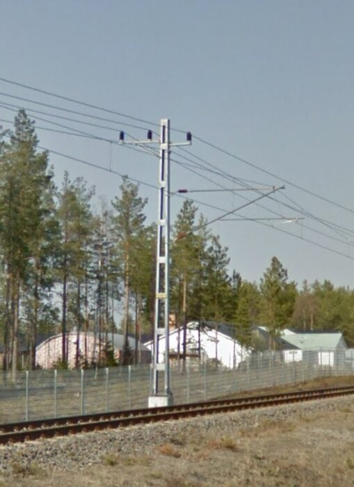 Järnvägsspår, stolpar, ledningar, träd, hus, klar himmel, dagtid.