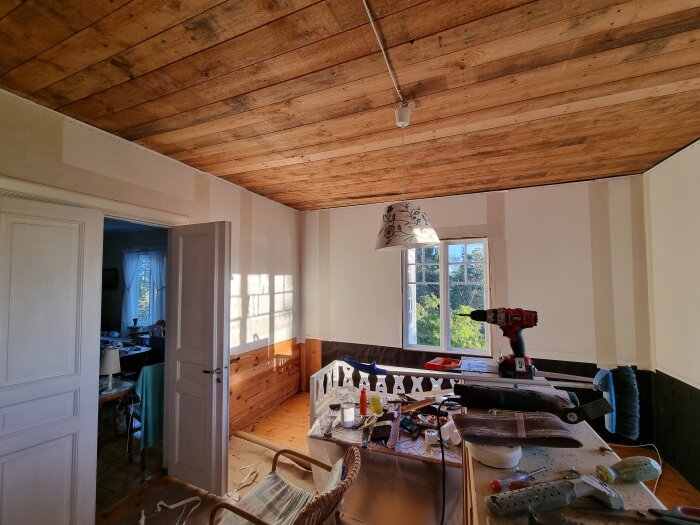 Rum under renovering, verktyg och målarutrustning utspridda, träbjälklag i tak, vitmålade väggar, öppen dörr till intilliggande rum.
