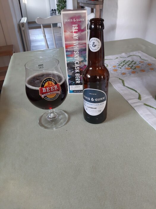 Glas med mörk öl, ölflaska och låda på bordet. Inredning, hemmiljö, Islay whisky cask beer.