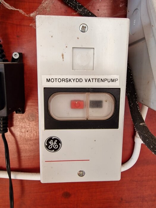 Äldre GE motorströmbrytare, etikett "MOTORSKYDD VATTENPUMP", smutsig, röd knapp på, röd bakgrund.