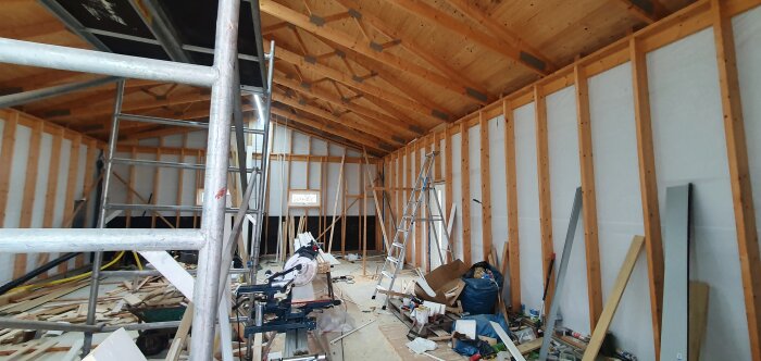 Inomhusbyggnadsplats med trästomme, isolering, rör, byggställningar och verktyg utspridda. Under konstruktion.