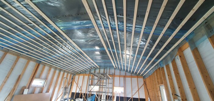 Pågående byggarbete, isolering och reglar i taket, osedda väggar, byggmaterial och ställning syns.