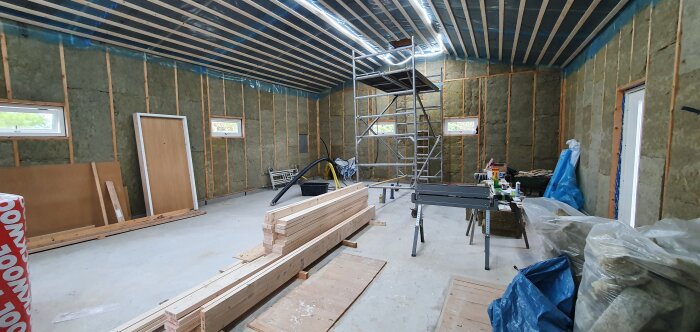 Byggarbetsplats inomhus med isolering, konstruktionsmaterial, verktyg och ställningar; pågående renovering eller uppbyggnad.