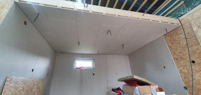 Ett rum under konstruktion med oslipade gipsskivor, synliga träbjälkar och isolering samt kartonger och en stege.