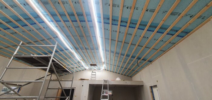 Inomhusbyggarbetsplats med isolering, träreglar i taket, byggställningar och belysning, under pågående renovering eller uppbyggnad.