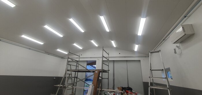 Industrilokal med vita LED-lampor, byggnadsställning, luftkonditionering och grått nedersegment på väggen.