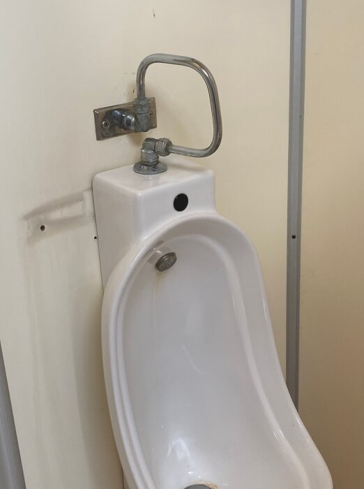 Keramiskt urinal med metallrör, väggmonterad spolmekanism, i en toalettstall.