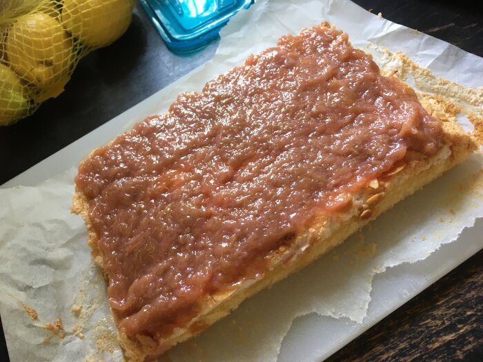 Bröd med rosa pålägg på bakplåtspapper, köksbänk, citroner i nät bakom.